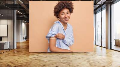 Pretty pleased Afr American woman in blue shirt raises sleeve shows vaccinated arm wears adhesive plaster smiles pleasantly isolated over beige background. Covid 19 protection and immunization Wall mural