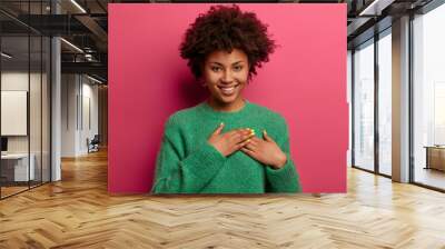 Pretty touched woman presses palms to heart, expresses positive feelings, feels touched to get help, makes gratitude gesture, wears warm green sweater, smiles sincerely, isolated on pink wall Wall mural