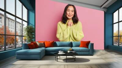 Pretty young Asian woman keeps hands together smiles pleasantly glad to receive compliment from boyfriend dressd in knitted yellow sweater isolated on pink background. Pleasant human emotions concept Wall mural