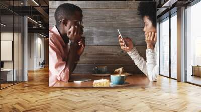 Profile of young dark-skinned couple sitting at restaurant: man looking bored and frustrated while his phone addicted pretty girlfriend browsing Internet on smart phone with happy face expression Wall mural