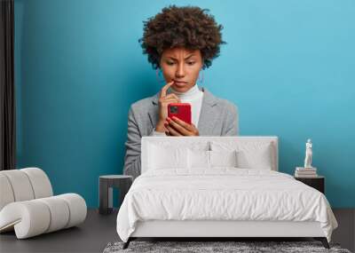 Puzzled confused dark skinned woman in formal wear looks indignant at smartphone, gazes at display, reads business news on website, isolated blue background. People and modern technology concept Wall mural