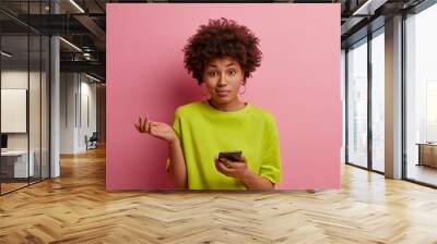 Puzzled dark skinned teenage girl shrugs shoulders, cannot understand how to use new application on mobile phone, uses modern device for communication, browses web, isolated on pink background Wall mural