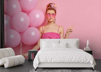 Puzzled redhead young woman dressed in glamour outfit wears everything pink poses on hens party with inflated balloons points at empty space bites lips shows place for your advertising content Wall mural