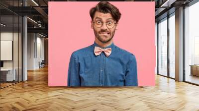 Puzzled shocked bearded young male bites lower lip, has trendy haircut and bristle, looks surprisingly at camera, wears fashionable shirt with pink bowtie. People, reaction and feelings concept Wall mural