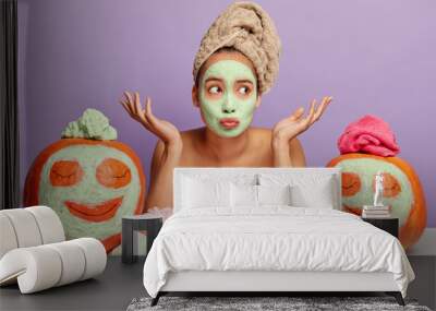 Puzzled unaware young woman with applied facial mask, spreads palms sideways, wears wrapped towel on head, uses cosmetics for beauty treatments, sits against purple wall with two orange pumpkins Wall mural