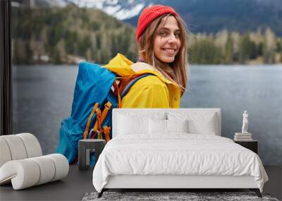 Rear view of happy young European woman enjoys nice serene day, nature landscape scenery, carries rucksack, stands sideways, wears red hat and yellow raincoat, spends free time in mountains. Wall mural