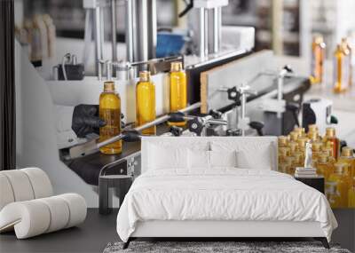 Rear view of unrecognizable factory worker in gown and gloves controlling production process of manufacturing cosmetics at modern laboratory, taking bottle of golden yellow liquid from conveyor line Wall mural