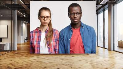 Relationships problems. Serious and sad young European female and Afro American male feeling unhappy after quarrel, standing against grey studio wall background, both wearing stylish eyeglasses Wall mural