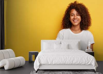 Relaxed female model with curly Afro hairstyle, grins from happiness, glad to have successful day, looks straightly at camera, wears white t shirt, laughs indoor over yellow background, copy space Wall mural