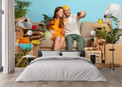 Relocating and changing house concept. Happy newlywed couple move to new abode, pose on cozy couch, glad live together, make selfie portrait on modern smart phone, empty room with packages around. Wall mural