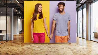 Romantic couple in love have date, hold hands, look positively at each other, feel support, walk together. Positive man poses over purple background, woman on yellow. Contrast. Relationship concept Wall mural