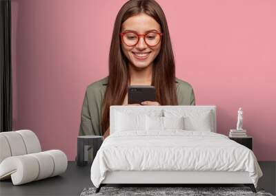 Satisfied lovely woman holds modern cell phone, texts with friend online in chat, dressed in stylish clothes, poses against pink wall, connected to high speed internet. Shot of teenager with device Wall mural