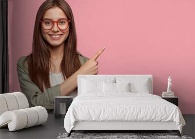 Satisfied young female with positive expression, attracts your attention at copy space aside, demonstrates some items in shop, dressed in casual shirt, has dark straight hair, isolated over pink wall Wall mural