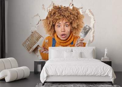 Scared curly female plasterer busy repairing wall in apartment holds brush and spatula being gypsum master holds breath from amazement poses in hole wears orange jumper. Home renovating concept Wall mural