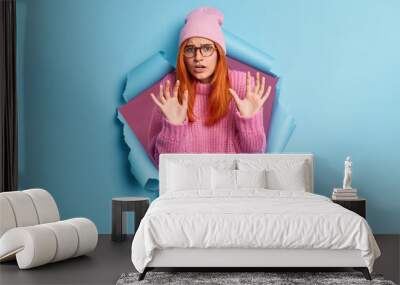 Scared fearful woman raises hands in defensive gesture sees something horrible wears pink hat and sweater breaks through paper background expresses negative emotions. People and fright concept Wall mural