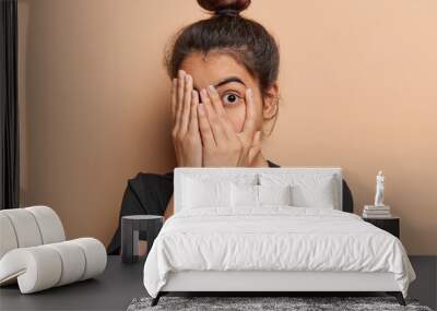 Scared Latin woman with hair bun peeks through her fingers fearfully hiding her face unable to bear sight of something terrible witnessing something truly terrible dressed in casual t shirt isolated Wall mural