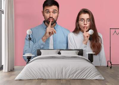 Secret and silence concept. Shot of surprised Caucasian lady and guy hold fore fingers over lips, make hush sign, dressed in casual clothes, isolated over pink background, tell secret to friends, Wall mural