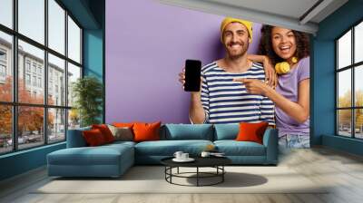 Selective focus of happy woman and man points to modern device with blank screen for your advertising, promot cool gadget, have smiles, wear stylish outfit, stands over purple wall, free space on left Wall mural