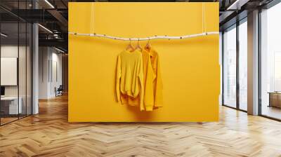 Selective focus. Three items of clothes on hangers. Long sleeved yellow jumpers on wooden rack near bright vivid wall. Copy space for text. Various casual outfits hanging in row at dressing room Wall mural