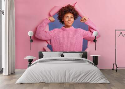 Self assured beautiful young Afro American woman raises arms shows biceps being strong and powerful proud of her own achievements wears casual knitted sweater breaks through paper background Wall mural