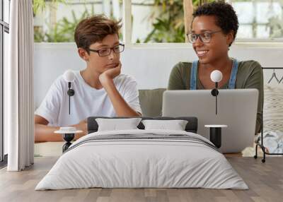 Self education and e learning concept. Satisfied black female volunteer tries to explain her strategy to young learner, watch attentively video in laptop computer. Tutor teaches skilled student Wall mural