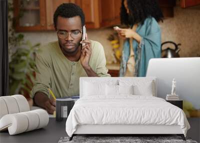 Serious African man having phone conversation with bank asking to extend loan term for paying out mortgage, holding pencil in the other hand, making notes in documents, lying on table in front of him Wall mural