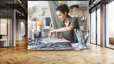 Serious brunette young beautiful woman sitting in art studio, taking colorful paints from tube while creating great masterpiece on easel, being preoccupied with her work, having nice imagination Wall mural