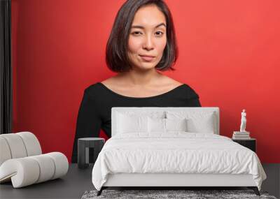 Serious confident young Asian woman keeps hands together has curious intention to do something looks attentively at camera wears black dress isolated over red background plans actions for future Wall mural