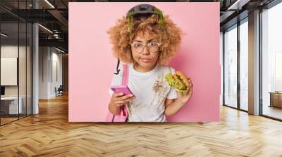 Serious curly haired woman focused into camera eats burger uses smartphone prefers fast food wears protective helmet and casual white t shirt isolated over pink background. Unhealthy eating. Wall mural