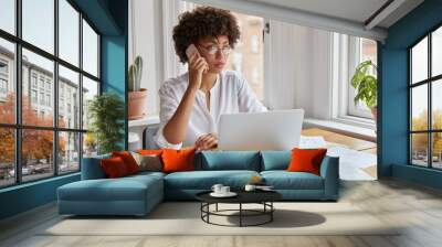 Serious ethnic female CEO wears round spectacles, tries to reach decision with colleague via cell phone, uses laptop computer, surrounded with papers, studies graphs, notices decrease in prices Wall mural