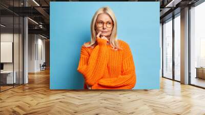 Serious European woman looks thoughtfully considers idea decides on something had doubts dressed in knitted orange jumper isolated over blue background thinks over dilemma. Face expressions concept Wall mural