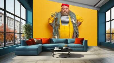 Serious fisherman looks with confident expression, keeps hands on hips, wears striped sailor shirt and yellow overalls, ready to catch fish with net, poses indoor. Fishery and hobby concept. Wall mural