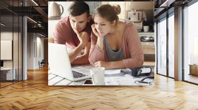 Serious man and woman sitting at kitchen table in front of open laptop computer, looking at screen with concentrated expression, focused on paying utility bills online. Family budget and finances Wall mural