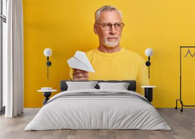 Serious thoughtful businessman concentrated somewhere holds paper plane as symbol of planning wears casual jumper and spectacles isolated over yellow background. Pensioner dreams about traveling Wall mural