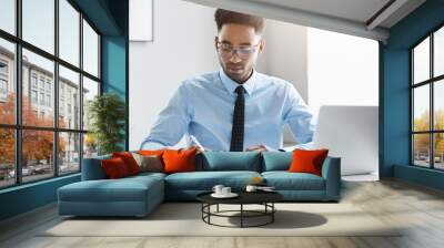 Serious unshaven dark-skinned businessman with curly dark hair, wearing glasses, elegant shirt and tie, writing with pen in his notebook, sitting at cozy cabinet, surrounded with modren gadgets Wall mural