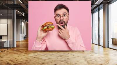 Shocked bearded adult man holds delicious hamburger eats fast food has unhealthy nutrition holds chin dressed in casual jumper isolated over pink background. Hungry guy with appetizing burger Wall mural