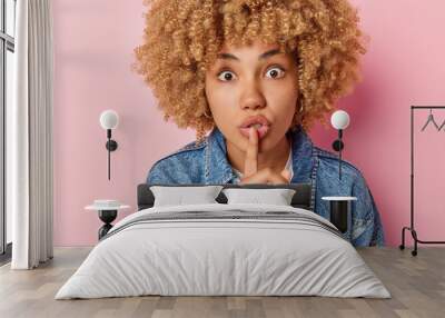 Shocked curly haired European woman stares bugged eyes presses index finger over lips shares secre with you wears denim jacket asks to be quiet and keep silence isolated over pink background Wall mural