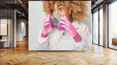 Shocked curly haired young woman wears respirator and protective suit liquidates agent protects herself from dangerous gases stares impressive away poses indoor afraids of chemical explosion Wall mural