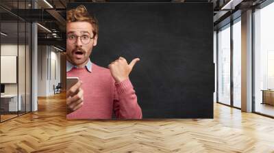 Shocked male student uses modern smart phone, messages with friend, notices something strange behind, isolated over black chalk background. People, technology, emotions and advertising concept Wall mural