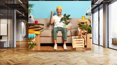 Shot of overjoyed guy clenches fist, poses at sofa, holds wrapped houseplant, moves in new modern house, being estate owner, has to unpack personal stuff from boxes. Home, tenant, relocation concept Wall mural