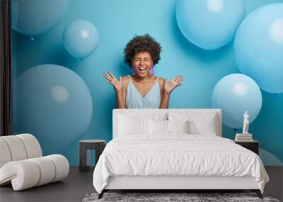 Shot of upbeat cheerful dark skinned woman feels very happy and excited, raises palms and laughs, spends free time on party, wears nice blue summer dress with earrings and rings, poses near balloons Wall mural