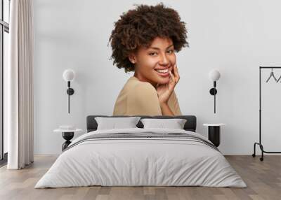 Sideways shot of cheerful dark skinned young model with Afro haircut, has gentle smile, dressed in casual beige sweater, models in studio against white background. People and ethnicity concept Wall mural