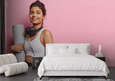 Sideways shot of Iranian woman dressed in sportswear holds foam roller has satisfied expression poses with headphones around neck isolated over pink background copy space for your promotional content Wall mural