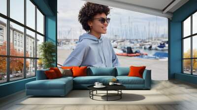 Sideways shot of positive curly haired woman poses on bench wears sunglasses and casual blue hoodie smiles broadly enjoys beautiful view of harbor breathes fresh marine air rests after walking Wall mural