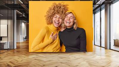 Sincere human emotions concept. Positive overjoyed young women have fun laugh gladfully smile toothily cannot stop laughing stand closely to each other dressed casually isolated over yellow wall Wall mural