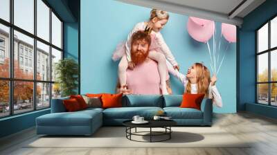 Sisterhood and festive day concept. Young man with tattoos, has thick ginger beard, gives piggy back to little girl, plays with sister, have overjoyed expressions, hold bunch of balloons, celebrate Wall mural