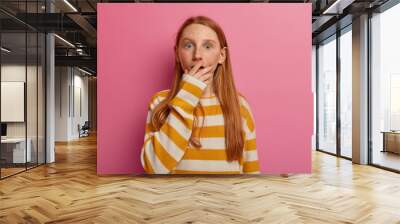 Small ginger girl covers mouth and stares horrified, being in panic as feels danger, got into trouble, looks with fear, wears striped jumper, isolated on pink background, wants to scream from fright Wall mural