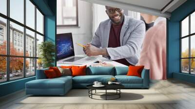 Smiling dark skinned male manager rejoices great success of his comapny, presents information to female partner, indicates at graphic on laptop computer, demonstrates application of new techniques Wall mural