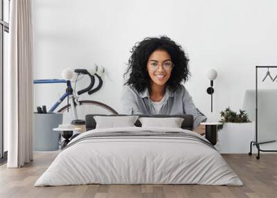 Smiling female student designer sitting at her workplace surrounded by gadgets being happy to finish her diploma paper and sketches. Pretty dark-skinned student working at drawings.Education concept Wall mural