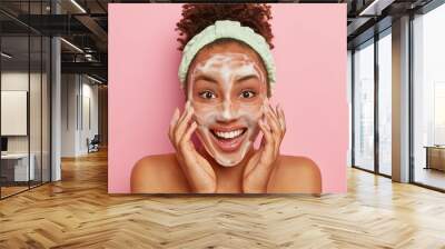 Smiling glad dark skinned young woman puts soap on face for cleansing, washes with cold water, wants to have healthy fresh skin., wears headband, looks positively at camera, stands naked indoor Wall mural